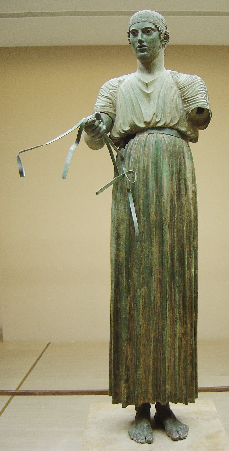 L’Aurige de Delphes, sculpture de Sculpteur grec anonyme : Jeune conducteur debout, vêtu d’une longue tunique plissée, au regard frontal et aux…