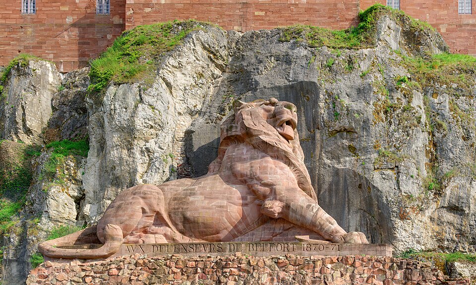 Le Lion de Belfort, sculpture de Auguste Bartholdi : Un lion monumental sculpté dans le grès rouge est couché, tourné vers la ville, dans une…