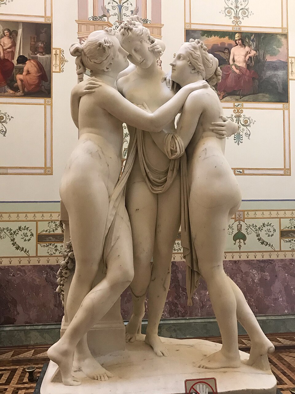 Les Trois Grâces, sculpture de Antonio Canova : Trois figures féminines nues s’enlacent autour d’un voile et d’un pilier, dans un marbre blanc…